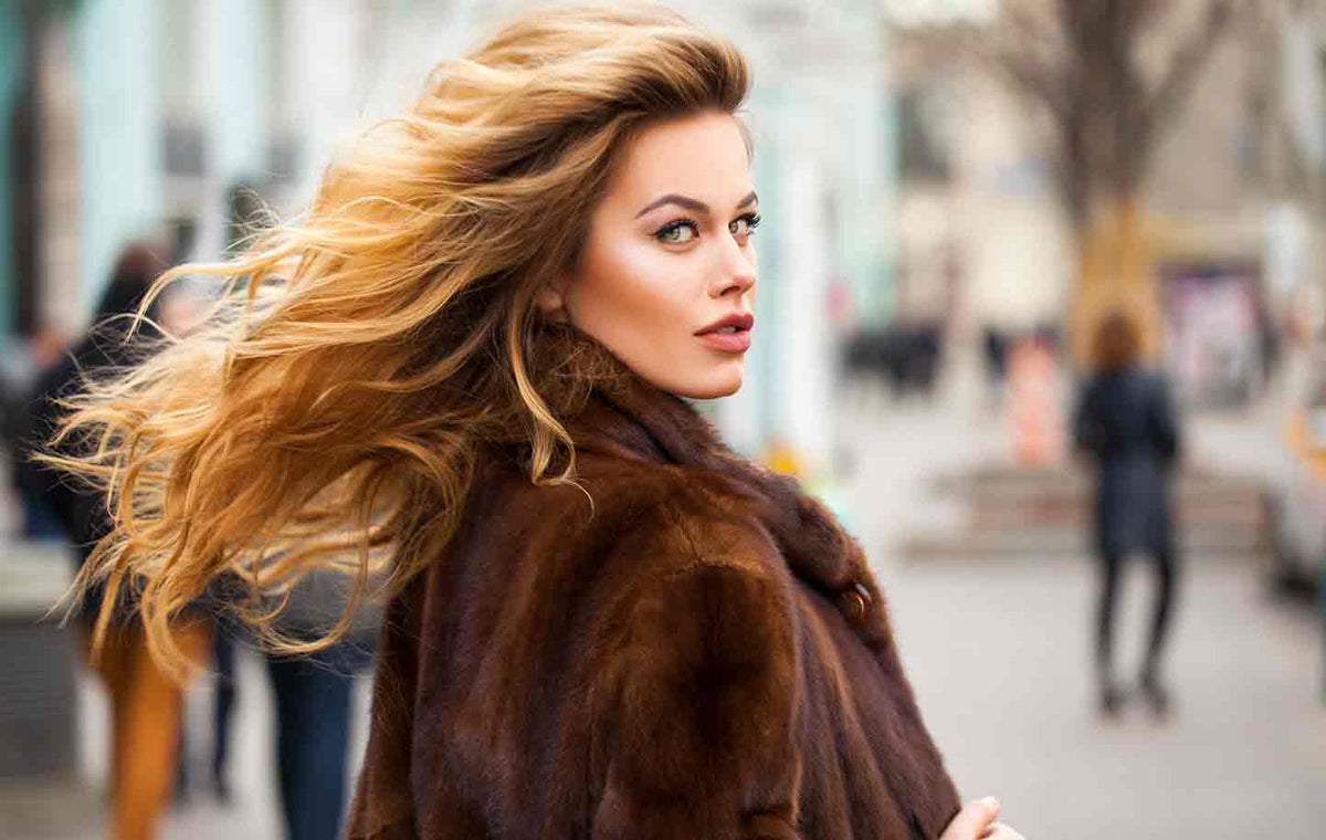 Close up of woman, hair blown back, wearing a winter fur coat against a blurred city background.