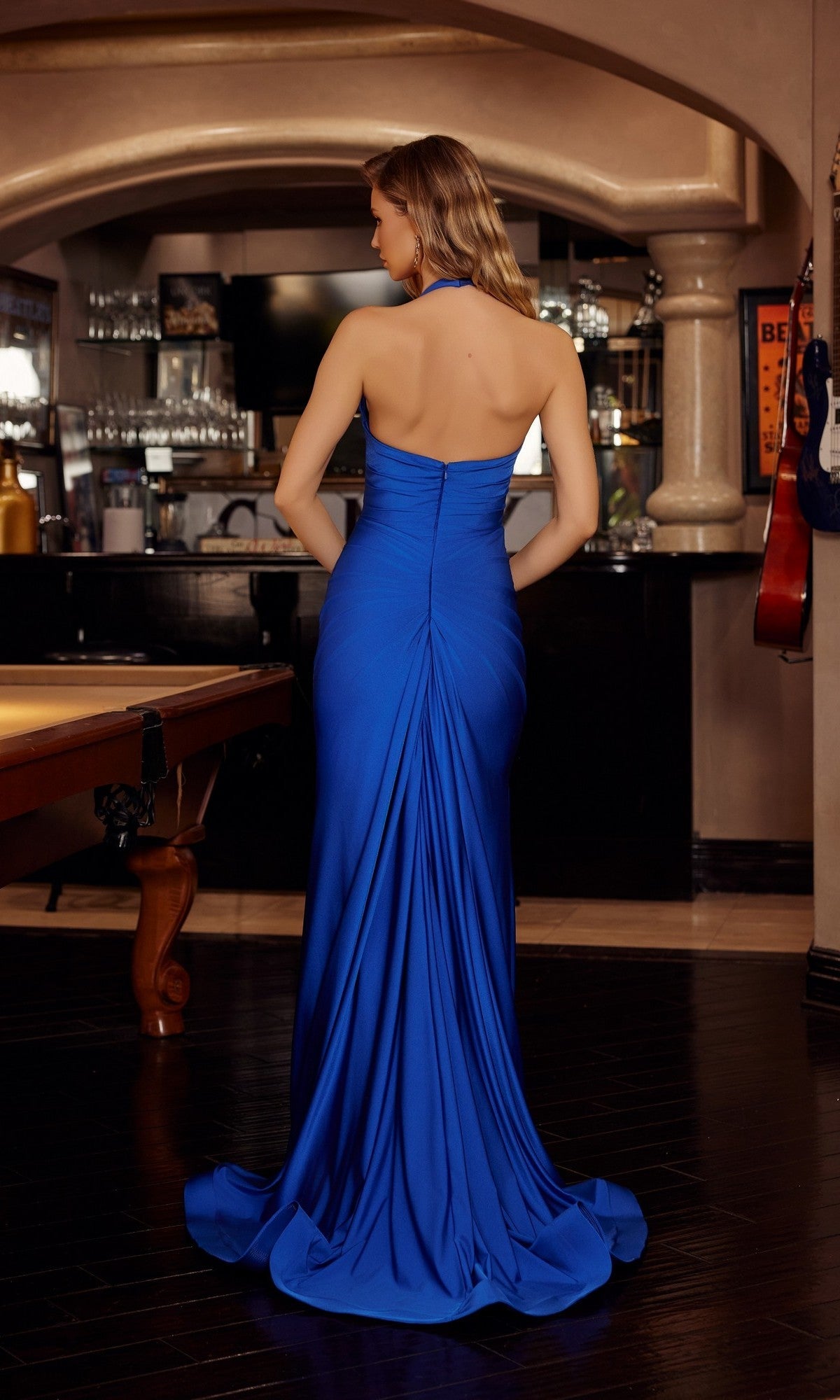 A woman in the Nox Anabel Long Prom Dress c1851, a vibrant blue halter gown by Nox Anabel, stands with her back to the camera in a room with a bar and pool table, ready for an unforgettable prom night. - Image