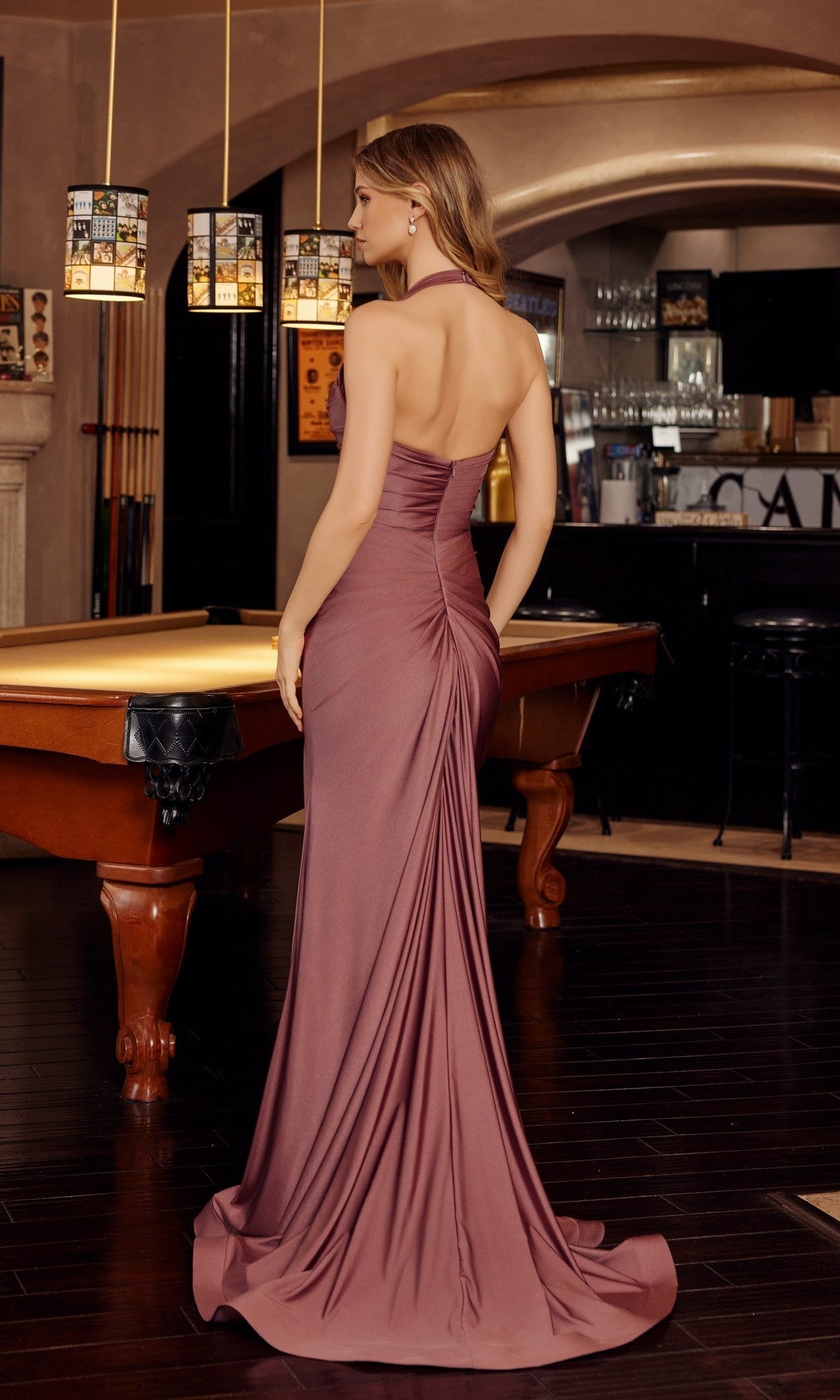 A woman in the Nox Anabel Long Prom Dress c1851, a mauve backless halter gown by Nox Anabel, stands by a pool table in a dimly lit room with dark wood floors and a bar, embodying the elegance of an unforgettable evening. - Image